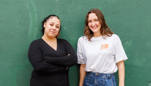 Two young women standing against green wall looking and smiling at camera